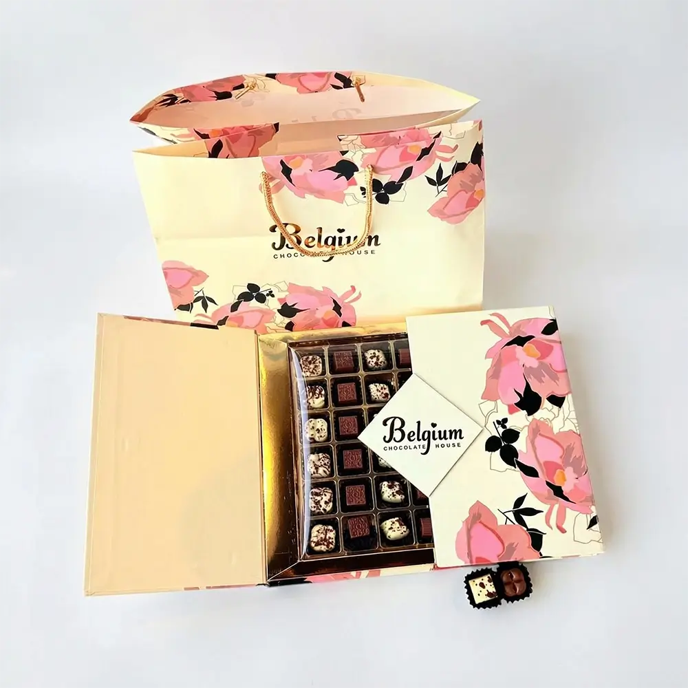 A floral-themed gift set featuring a pastel yellow box and matching bag labeled “Belgium Chocolate House.” The box is open to reveal an assortment of neatly arranged chocolates in different shapes and toppings, with a small branded card placed on top. The packaging is decorated with pink and black floral patterns, giving it an elegant, gift-ready appearance.