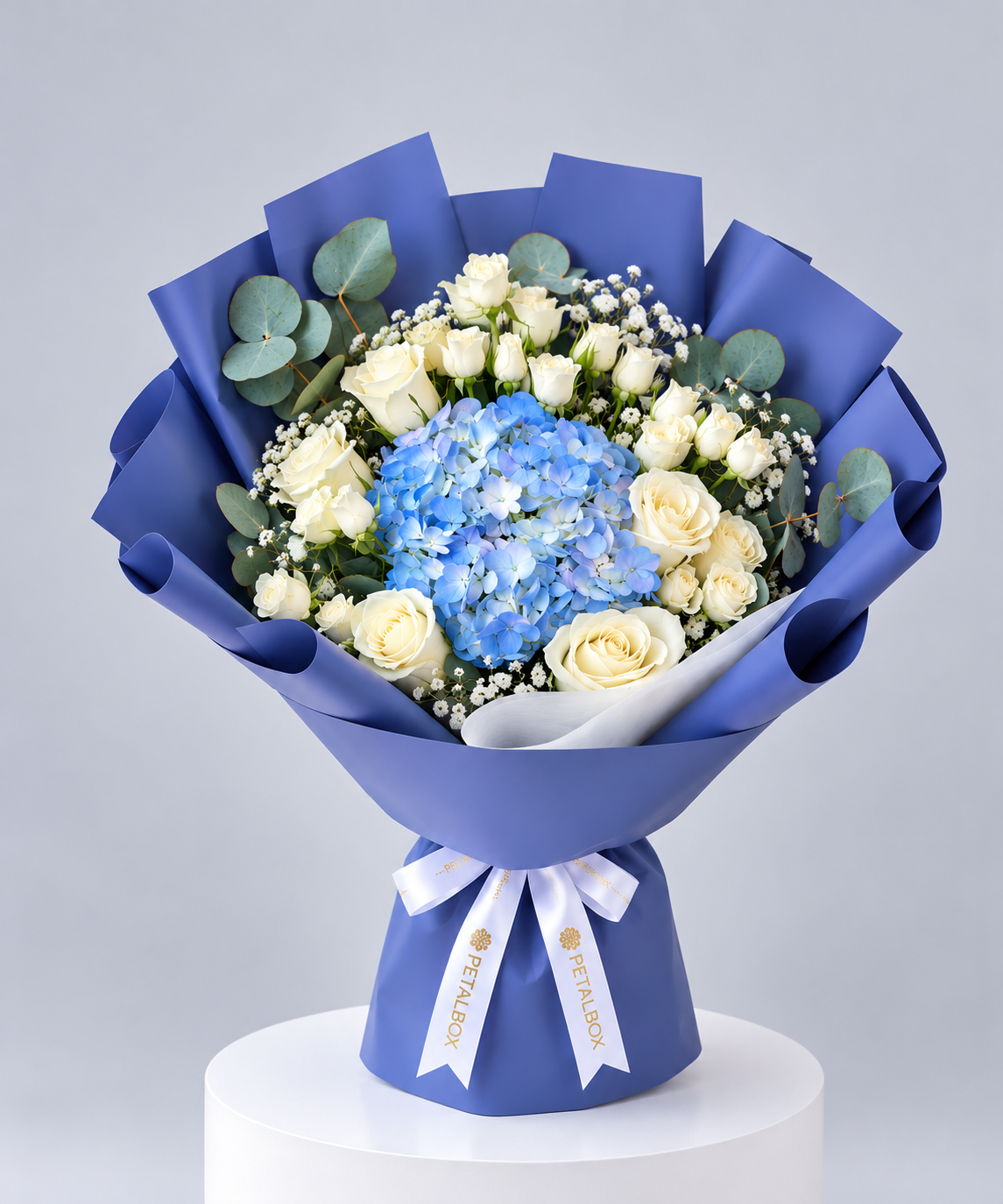Premium Petal Box bouquet featuring blue hydrangeas, ivory roses, delicate white baby’s breath, and eucalyptus foliage, wrapped in elegant blue paper with a white branded ribbon. Luxury flower delivery in Abu Dhabi and Dubai, UAE.