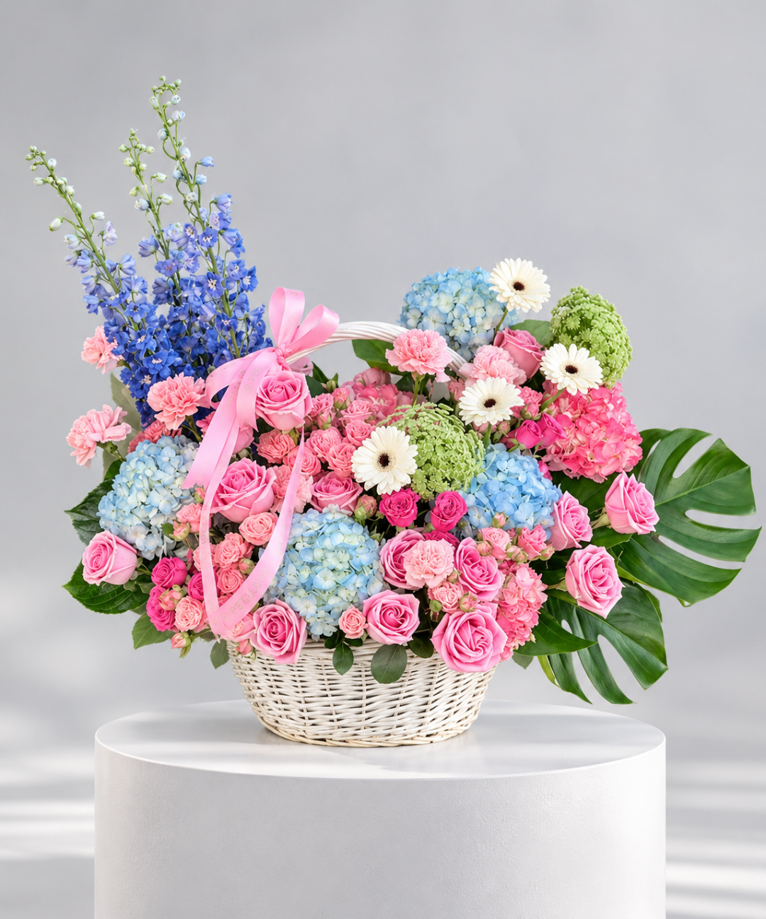 Luxury pastel flower basket featuring pink roses, carnations, blue delphiniums, hydrangeas and white gerbera daisies in a white wicker basket, ideal for gifting in Abu Dhabi and Dubai, UAE.