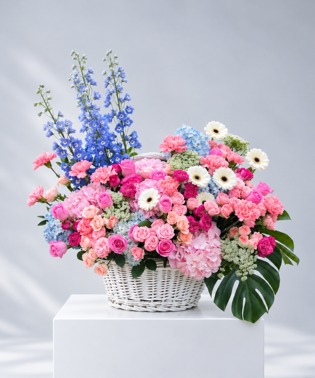 Luxury pastel flower basket featuring pink roses, carnations, blue delphiniums, hydrangeas and white gerbera daisies in a white wicker basket, ideal for gifting in Abu Dhabi and Dubai, UAE.
