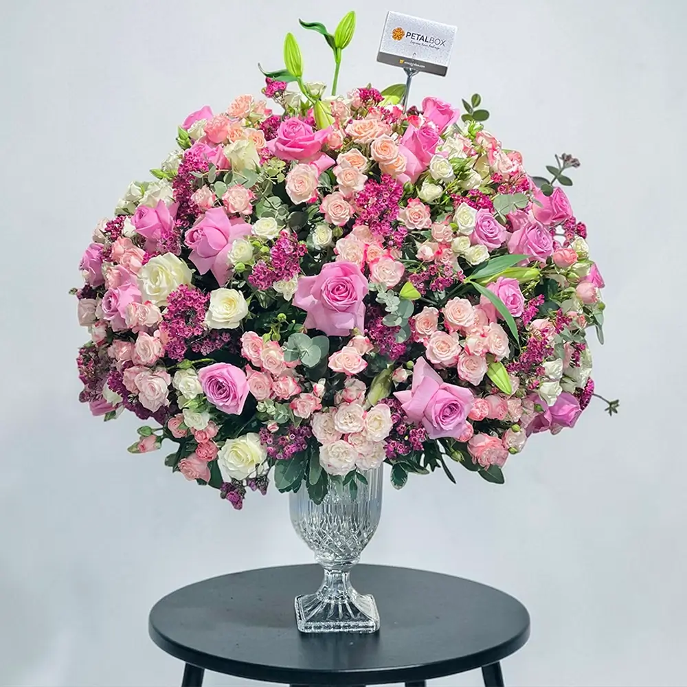 Luxury flower arrangement in a clear glass vase featuring pink and white roses, spray roses, lilies, purple filler flowers, and eucalyptus on a white background.