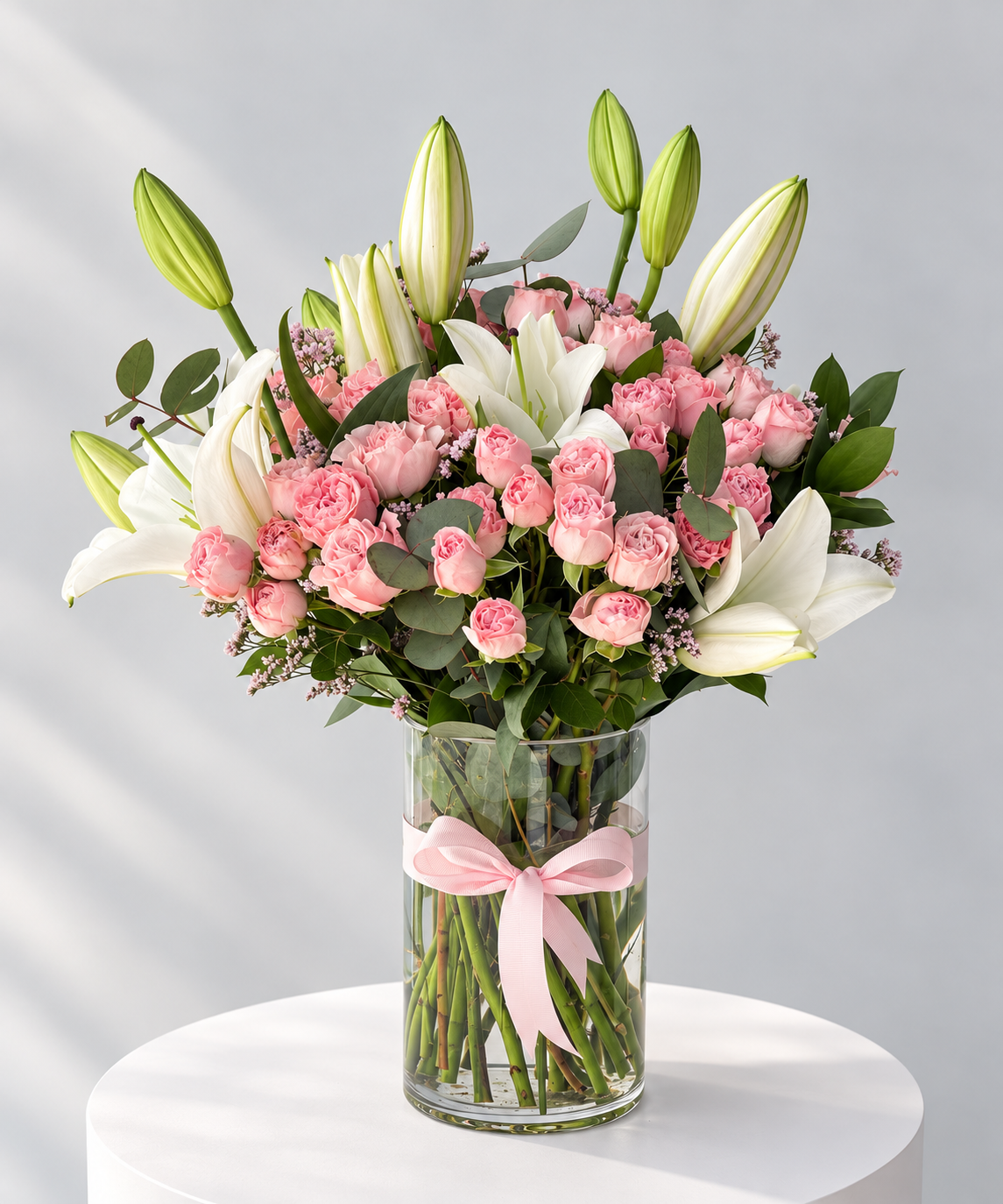 A floral bouquet in a clear glass vase with a pink ribbon, featuring white lilies, pink roses, and green buds on a white round pedestal against a light gray background.