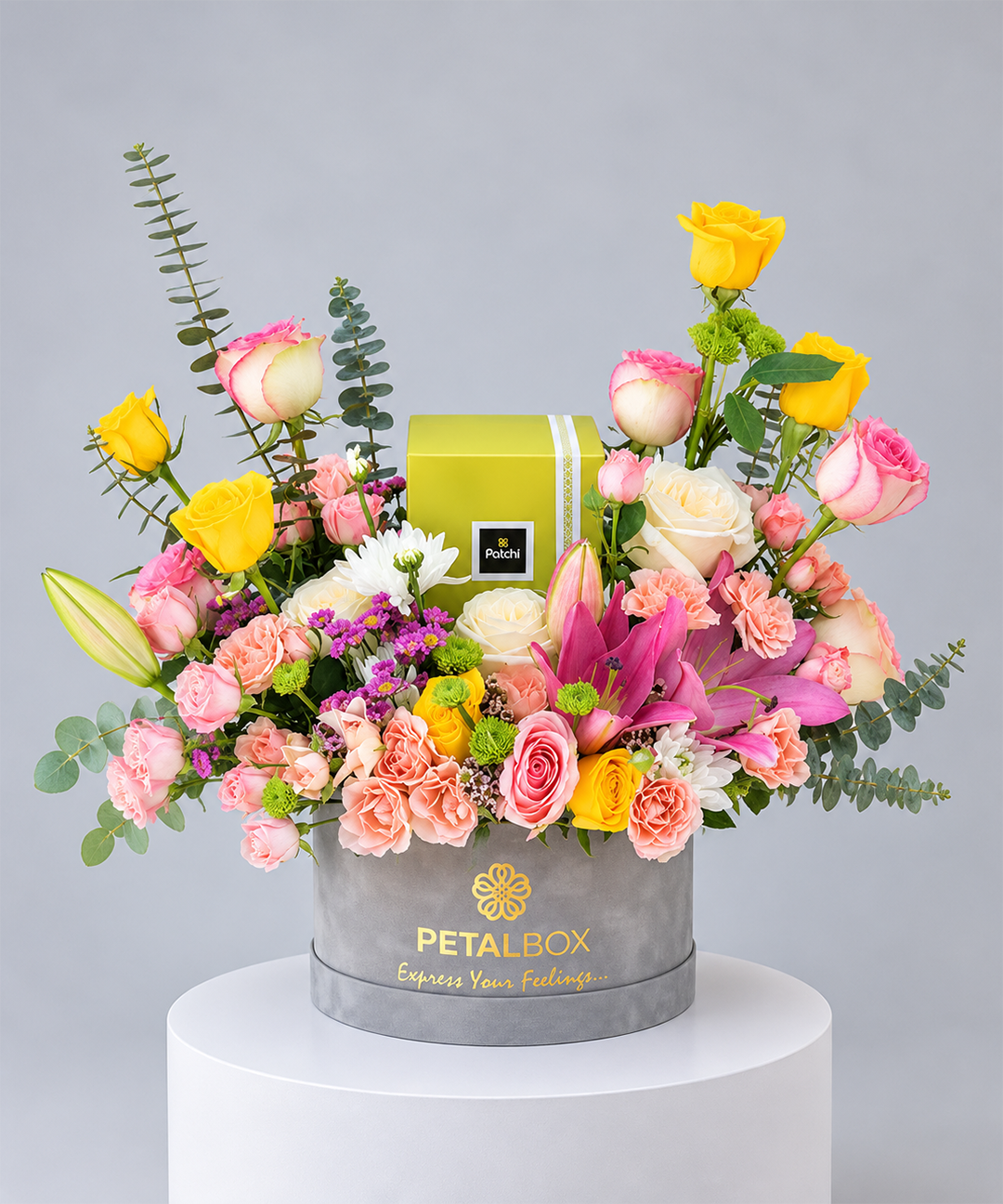 luxury mixed flower arrangement in a grey Petal Box hatbox featuring yellow roses, pink and cream roses, pink lilies, peach carnations, white chrysanthemums and eucalyptus foliage, paired with a Patchi chocolate box, premium flower gift for Abu Dhabi and Dubai