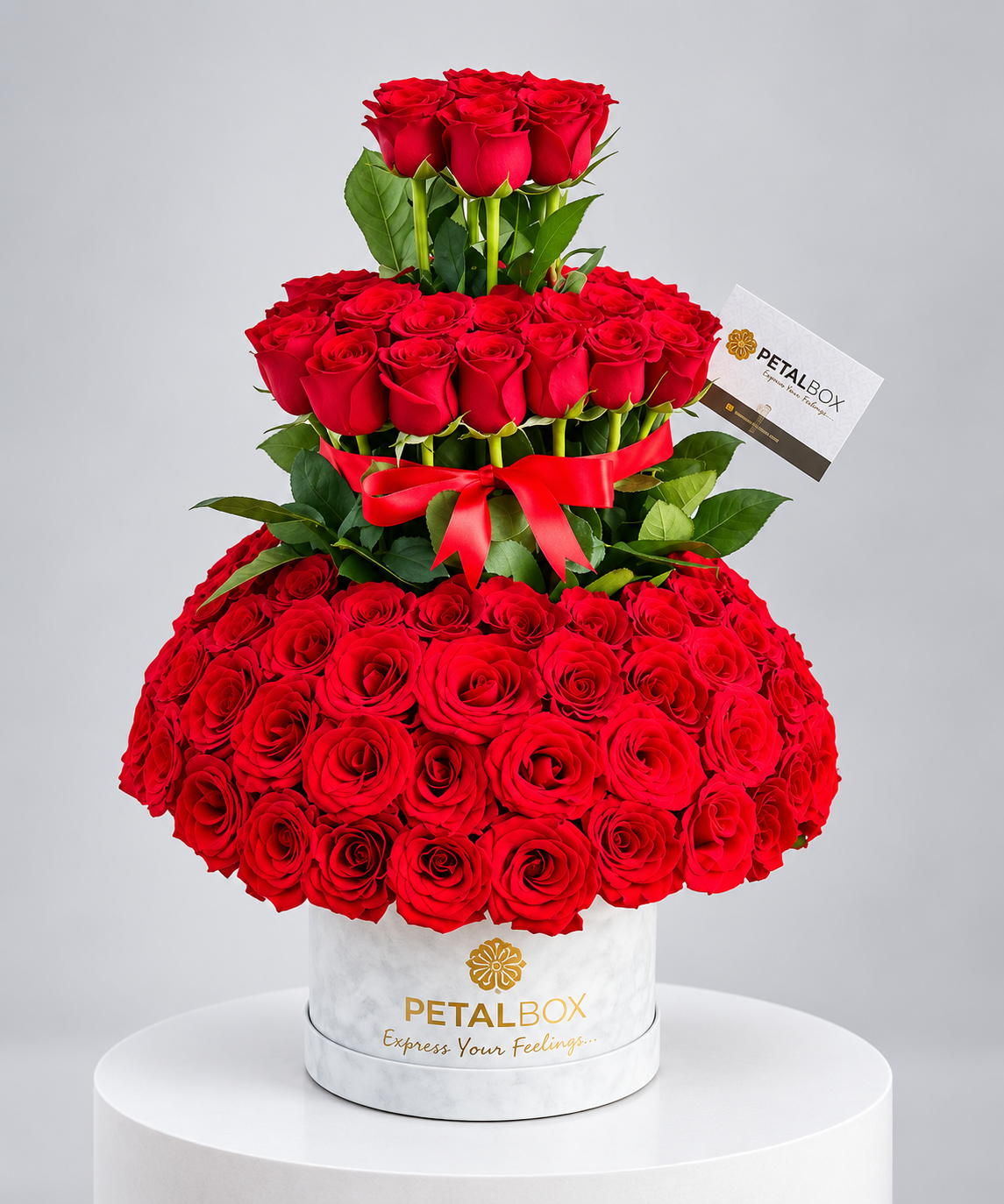 luxury tiered red rose arrangement featuring multiple layers of fresh red roses, lush green foliage and a red ribbon, displayed in a white marble Petal Box hatbox for premium gifting in Abu Dhabi and Dubai