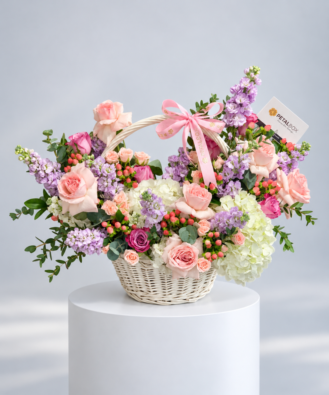 Purple and white stock, pink roses and white hydrangea in a white handled basket