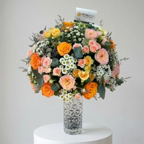 Elegant orange and blush rose vase arrangement featuring premium peach garden roses, orange roses, delicate spray roses, white daisy blooms, eucalyptus foliage, and seasonal fillers styled in a luxury crystal glass vase.