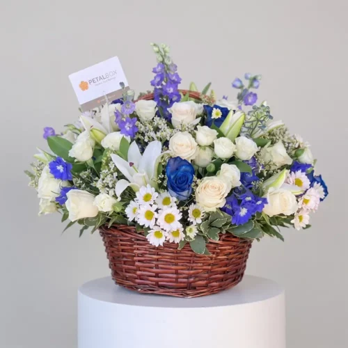 luxury blue and white flower basket with blue roses, white lilies, white roses, delphiniums and daisies in a woven basket, premium floral arrangement by Petal Box UAE for flower delivery in Abu Dhabi and Dubai