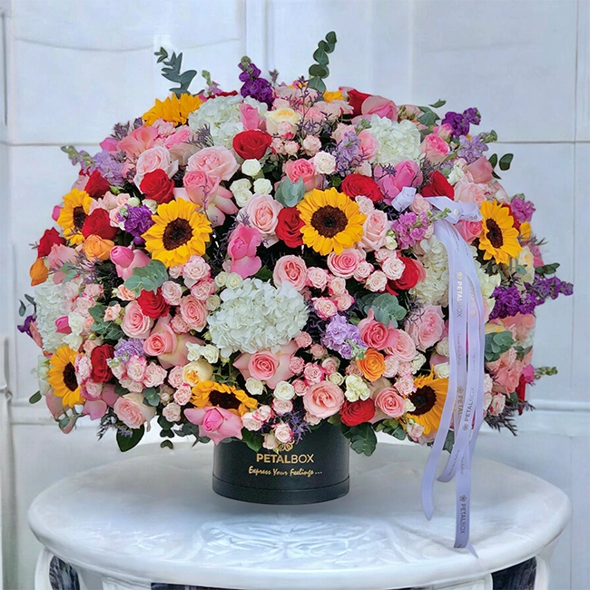 Luxury Petal Box flower arrangement in a black hat box featuring pink and red roses, sunflowers, white hydrangeas, pastel spray roses, and mixed seasonal blooms displayed on a marble table.