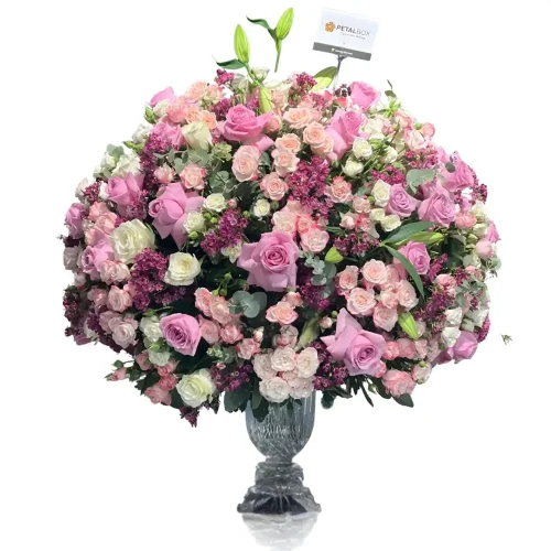 Luxury flower arrangement in a clear glass vase featuring pink and white roses, spray roses, lilies, purple filler flowers, and eucalyptus on a white background.