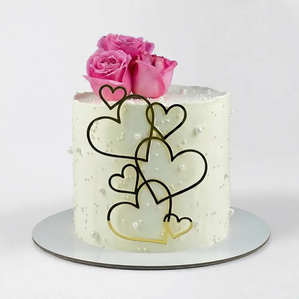 Minimal white cake with cascading gold heart outlines, fresh pink roses on top and pearl dot texture