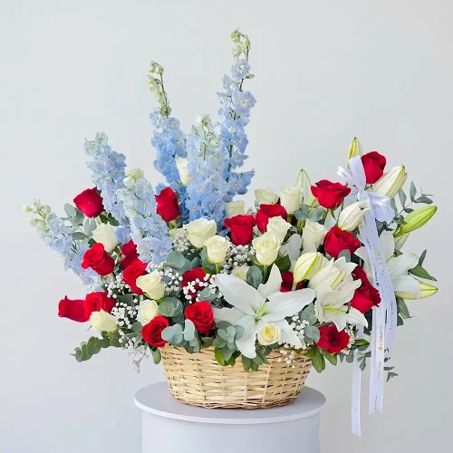 Mixed Flower Gift Basket by Petal Box with blue delphinium, white lilies, red and white roses, gypsophila, eucalyptus, and green leaves.